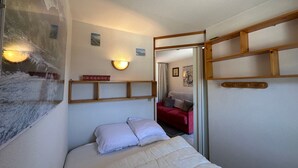 A bedroom with a bed, a wall-mounted shelf, a wall sconce, and a mirror reflecting a red sofa and a painting. - Cozy 2-room flat without lift, for 4/5 people, Morillon 1100 (Morillon 1100)