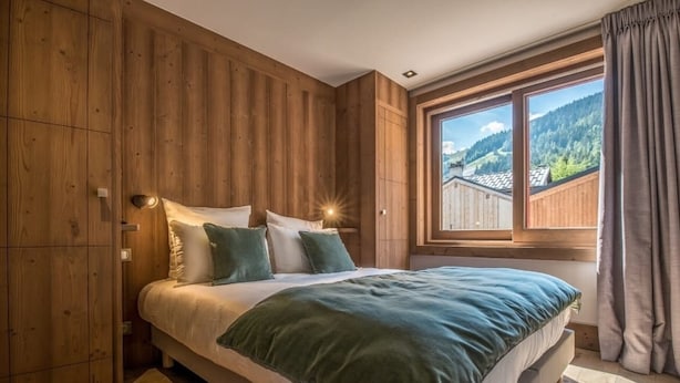 A bedroom with a wooden wall, a large bed with green bedding, and a window with a mountain view.
