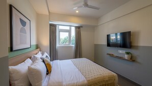 A hotel room with a bed, a ceiling fan, a TV mounted on the wall, and a window with curtains. - Olive Hotel JP Nagar by Embassy Group (Bengaluru)