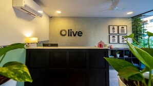 A reception desk with a 'live' sign, air conditioning, ceiling fan, and framed artwork on the wall. - Olive Hotel JP Nagar by Embassy Group (Bengaluru)
