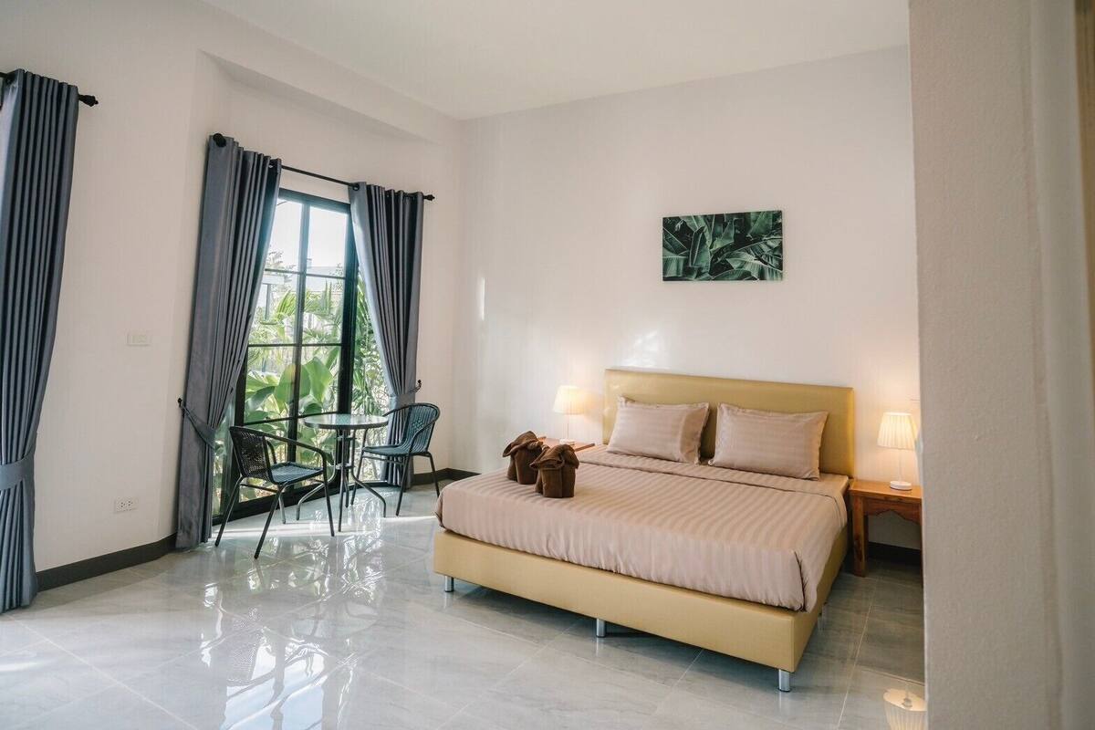 A modern hotel room with a large bed, a dining area with chairs, and a view of greenery outside.