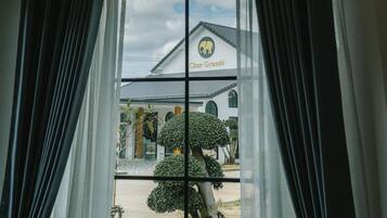 Vista a través de una ventana con cortinas, mostrando un edificio con un letrero que dice "Chor Grande" y un jardín cuidado con un árbol esférico.