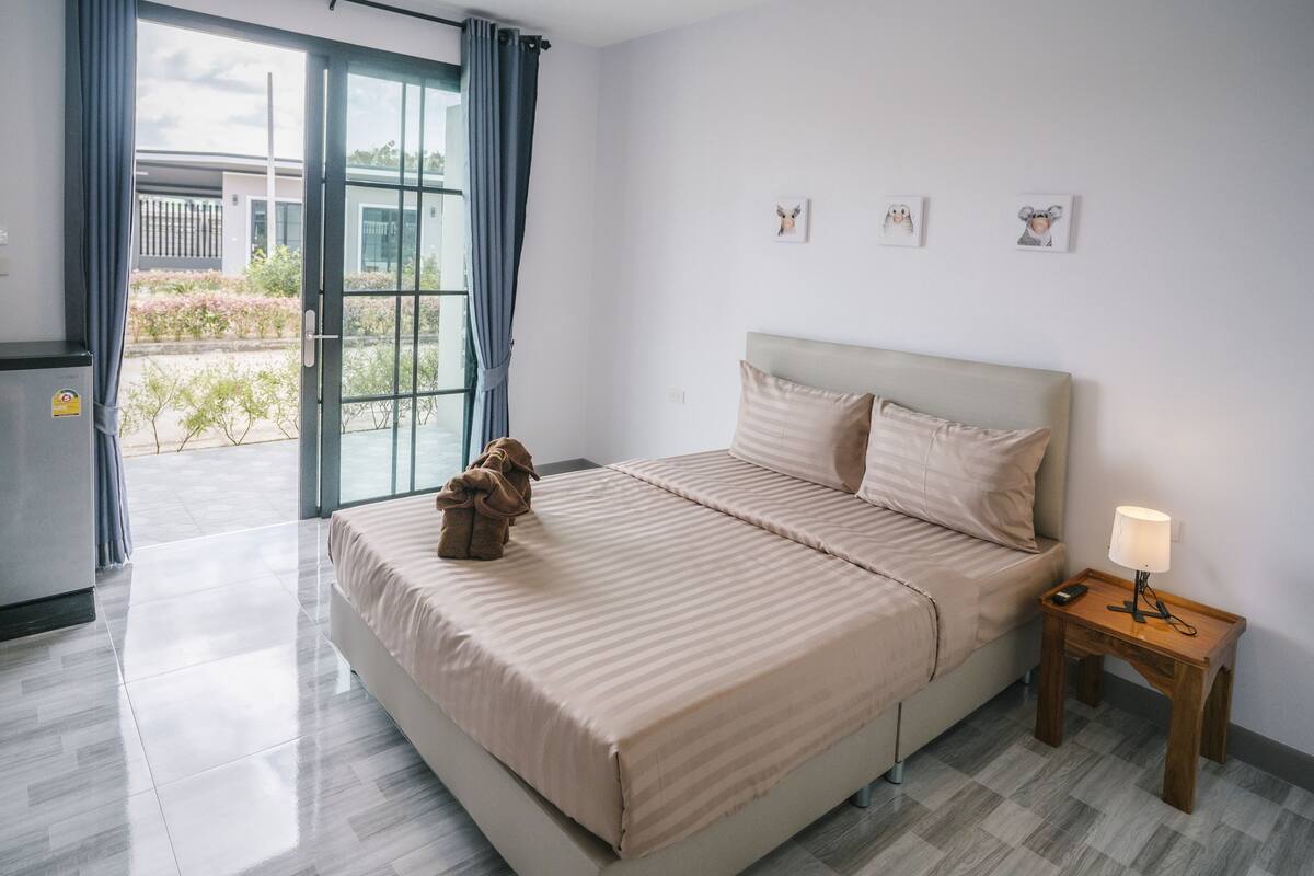 A modern hotel room with a large bed, a bedside table with a lamp, and a view of the outdoors through a sliding glass door.