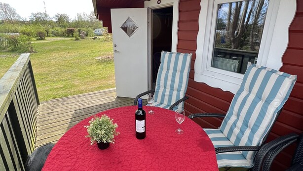 Un espace extérieur abrité, aménagé avec une table recouverte d’une nappe rouge, une bouteille de vin et deux chaises à rayures.