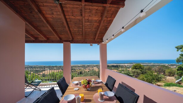 Mesa de madera preparada para el desayuno, con sillas, vista al mar y un balcón con barandilla.