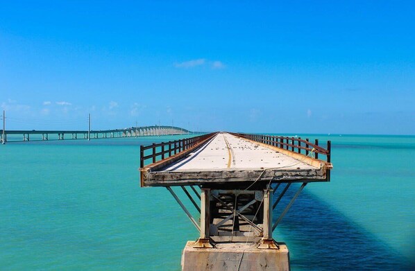 Un muelle que se adentra en un mar azul y transparente, con un puente distante.