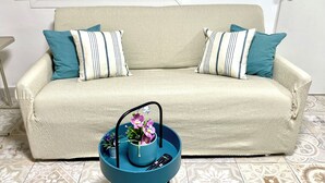 Un sofá beige con almohadas a rayas, una mesita auxiliar azul con un control remoto y flores, y baldosas de suelo estampadas.