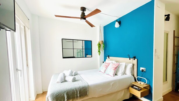 Un dormitorio con cama, ventilador de techo, mesita de noche con lámpara y toma de corriente en la pared.