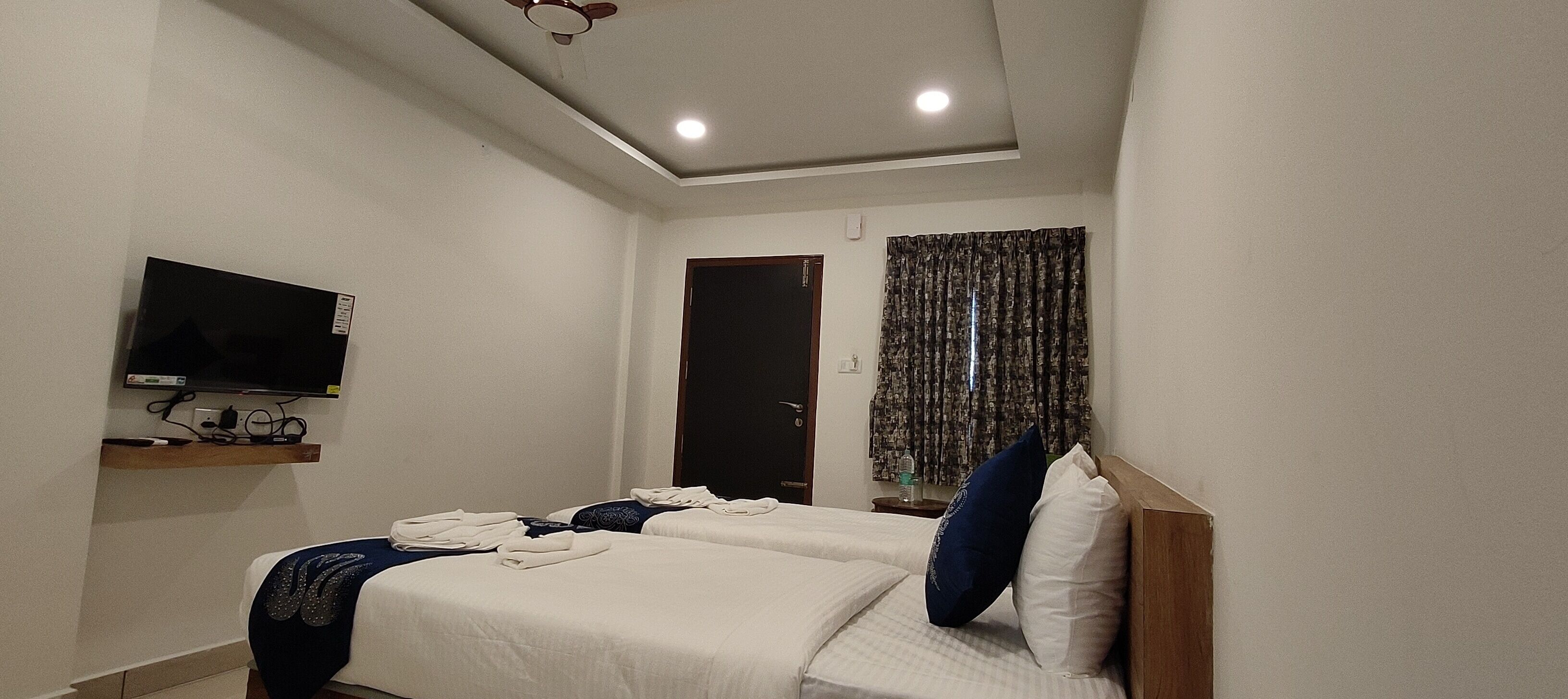 A hotel room with two beds, a flat-screen TV mounted on the wall, a ceiling fan, and a door with a curtain.