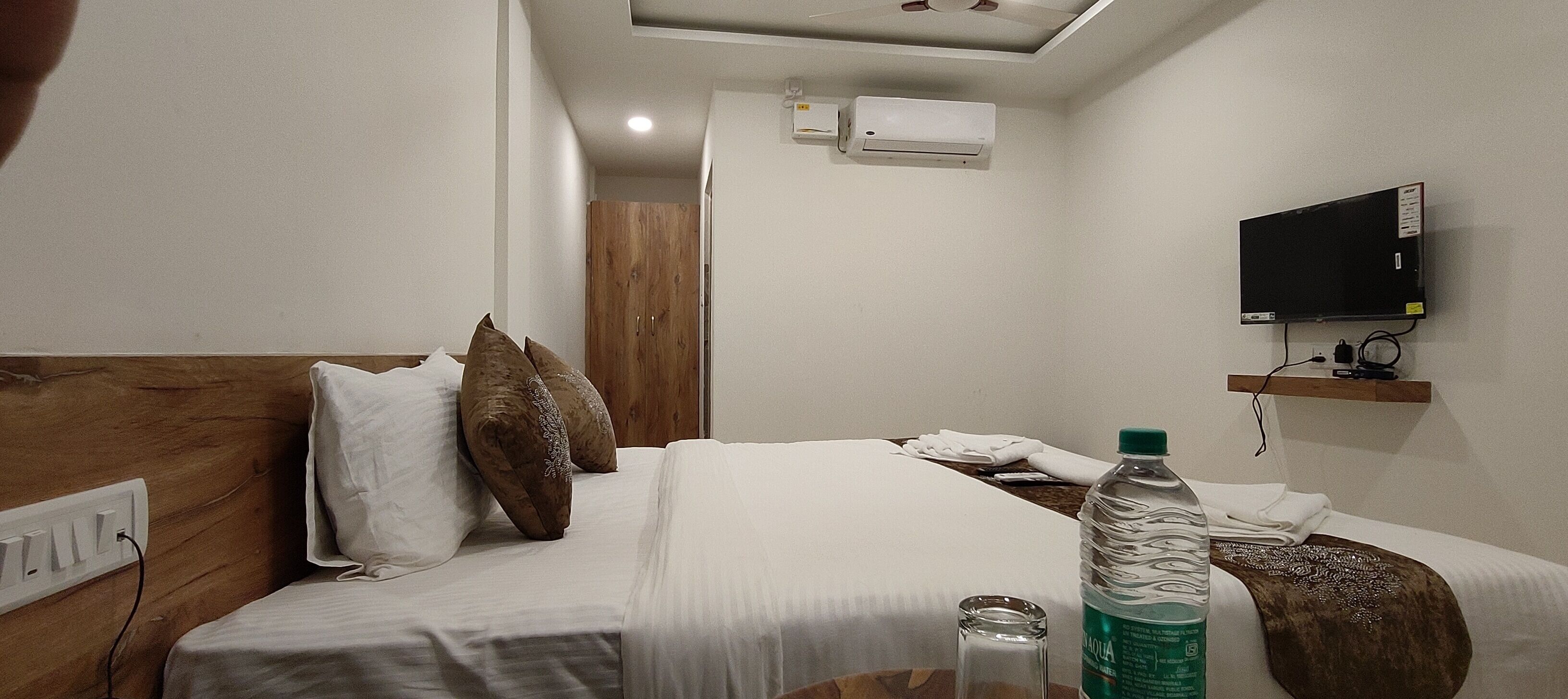Ein Hotelzimmer mit einem hölzernen Kopfteil, einem Bett mit weißer Bettwäsche, einer Wasserflasche, einem Fernseher auf einem Regal und einer Klimaanlage an der Wand.