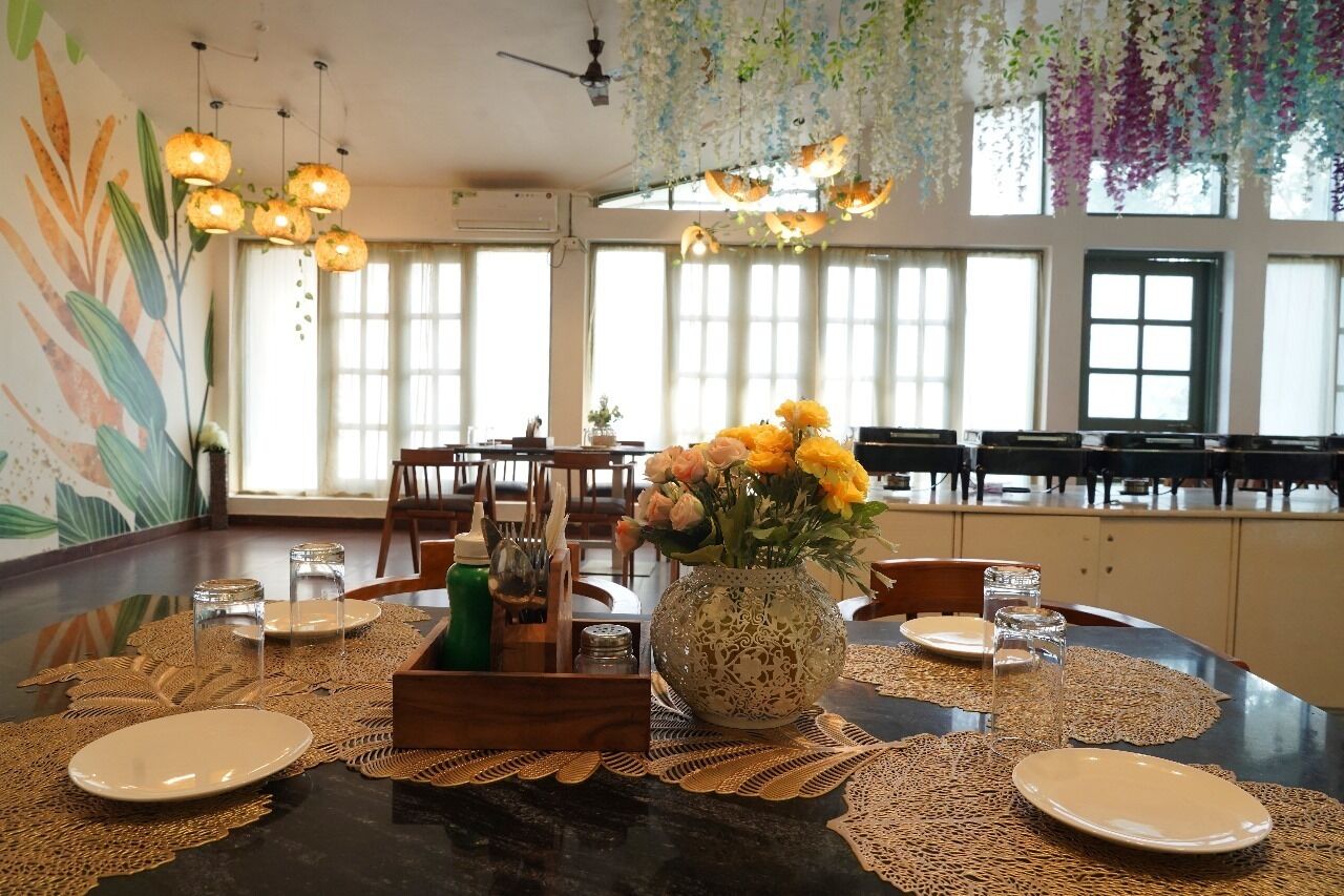 A dining area with a round table set for four, surrounded by chairs. A vase with yellow flowers is placed on the table, and there are placemats and cutlery.