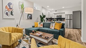 Un salon moderne avec un canapé d’angle, des fauteuils jaunes et une table basse. On aperçoit, au fond, une cuisine équipée d’appareils électroménagers en acier inoxydable.