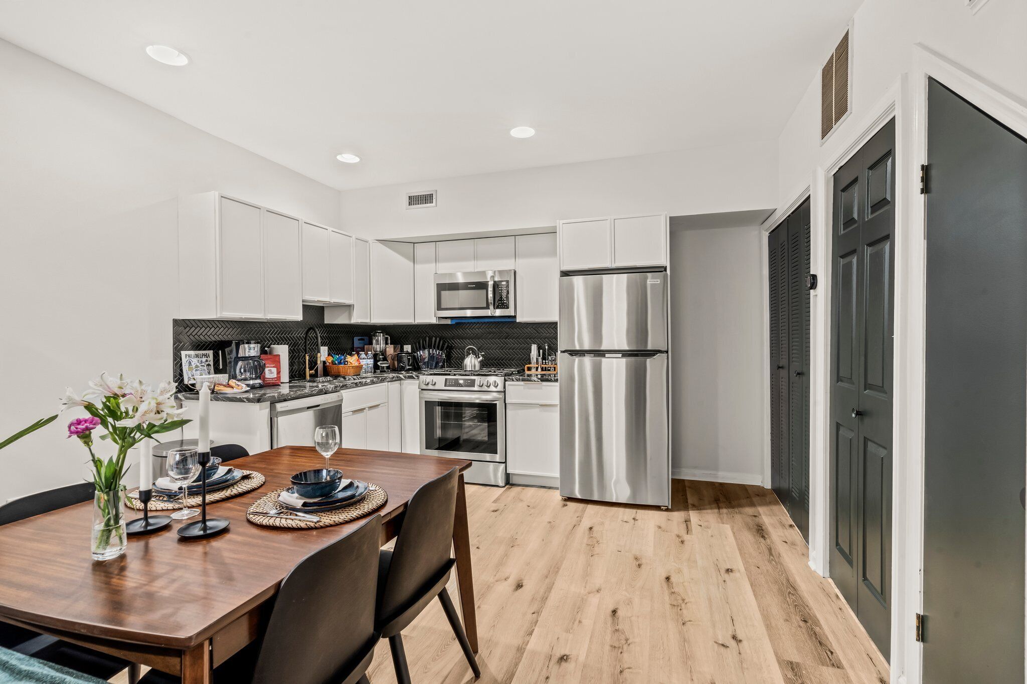 A modern kitchen with white cabinets, stainless steel appliances, and a wooden dining table set for two.