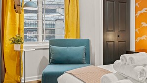 Une chambre d’hôtel avec un lit, une chaise bleue, une fenêtre avec des rideaux jaunes, une plante en pot et un mur orné d’un motif représentant un lion.