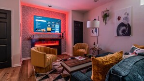 Un salon moderne avec un téléviseur à écran plat fixé sur un mur à motifs, une cheminée, deux fauteuils, une table basse et un canapé agrémenté de coussins décoratifs.