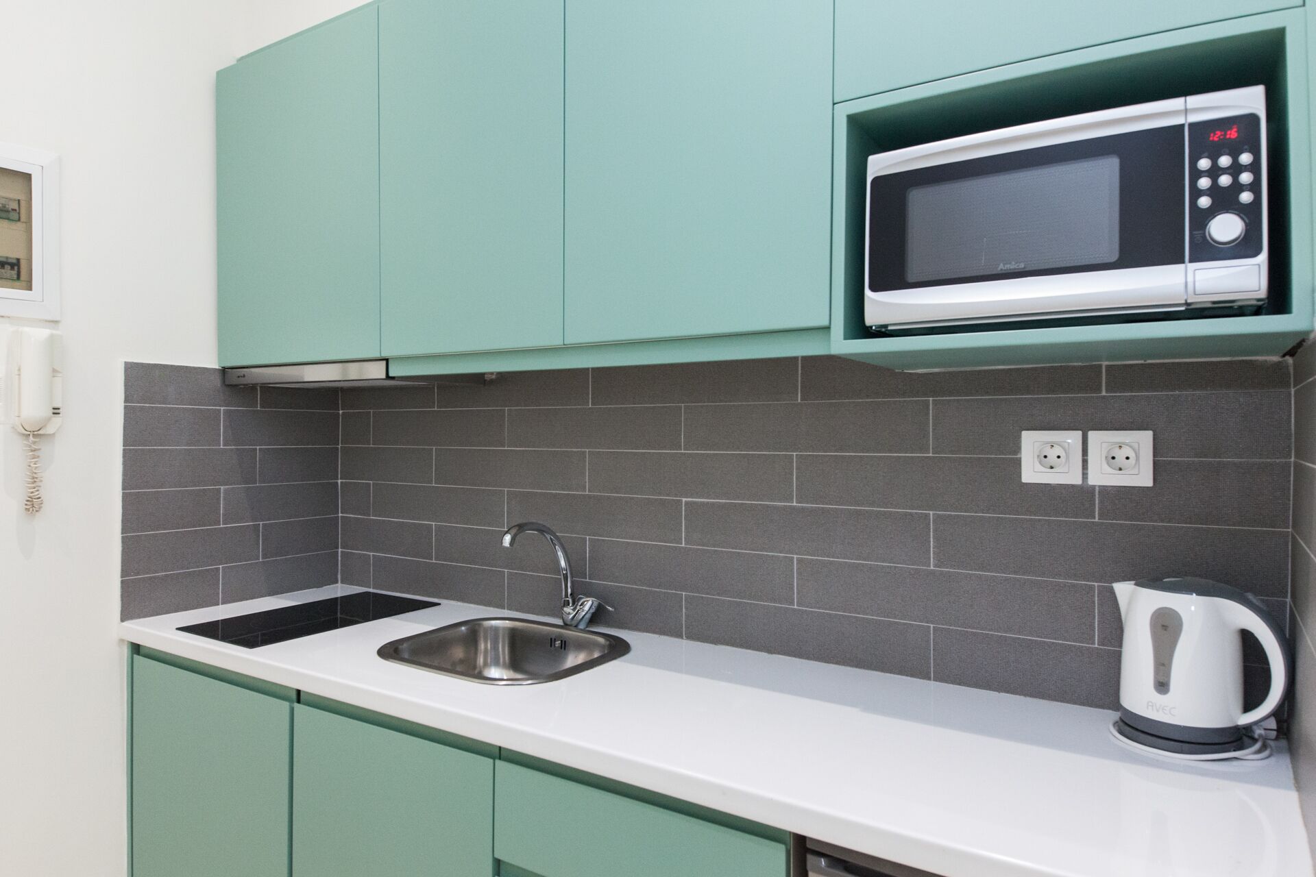 Een keuken met blauwgroene kastjes, een roestvrijstalen spoelbak, een magnetron en een waterkoker.
