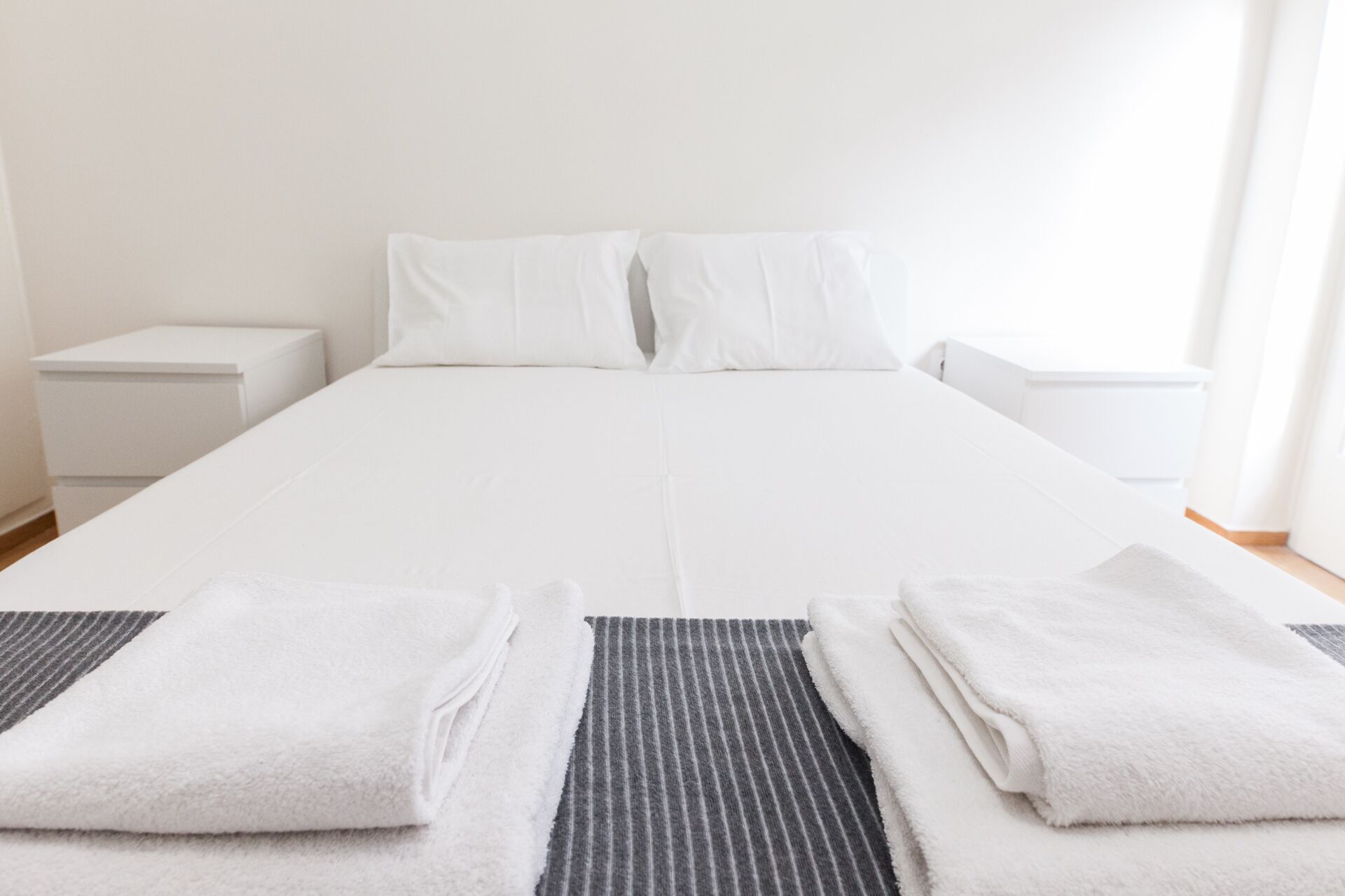 A neatly made bed with white linens, two pillows, and two folded towels on a striped rug.