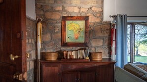 Un mur en pierre orné d’une carte de l’Afrique encadrée, un meuble en bois contenant des livres et des paniers, et une fenêtre avec des rideaux.
