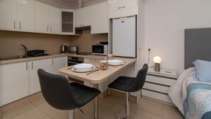 Cocina compacta con armarios blancos, microondas, hervidor, y refrigerador. Hay una pequeña mesa de comedor con dos sillas y una cama con manta azul.