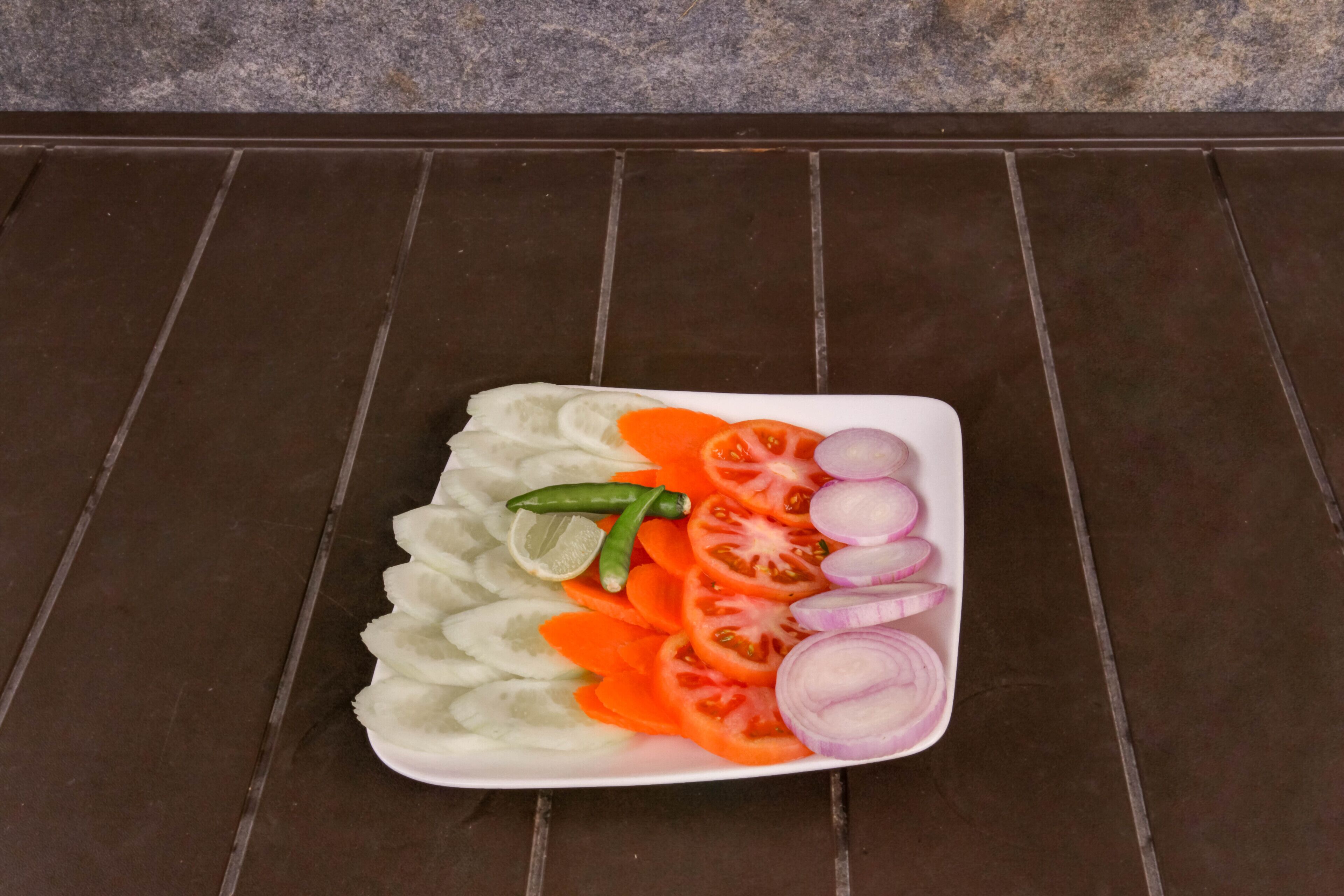 Een wit bord met plakjes komkommer, tomaten, wortels, rode uien en groene pepers op een donkere betegelde ondergrond.