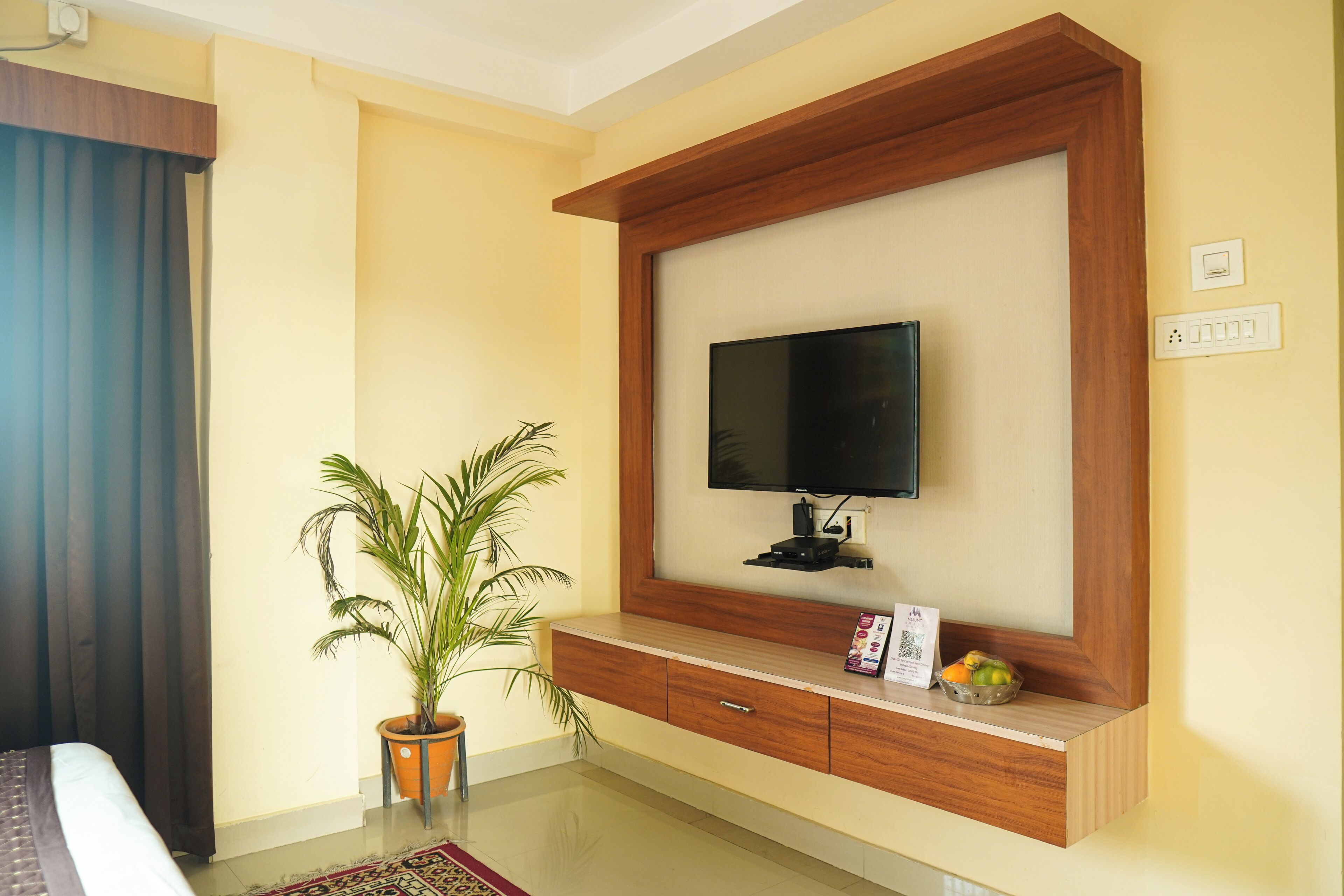 A hotel room with a wooden TV stand, a flat-screen TV, a potted plant, and a small table with a fruit bowl.