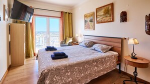 Ett hotellrum med en säng, en tv som är monterad på väggen, ett fönster med gardiner, en stol, ett nattduksbord och en lampa.