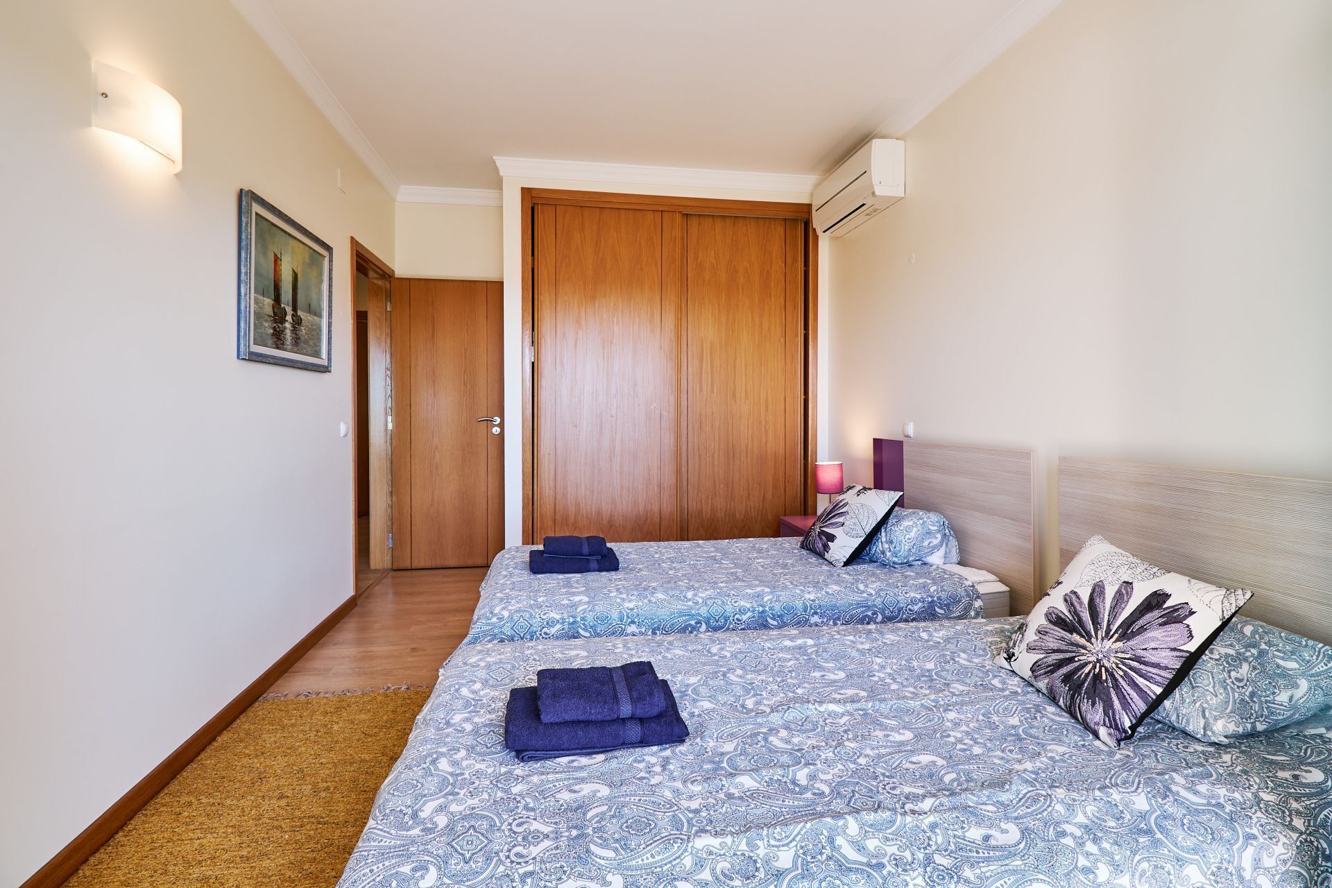 A hotel room with two beds, a wooden wardrobe, a painting on the wall, and a bedside lamp.