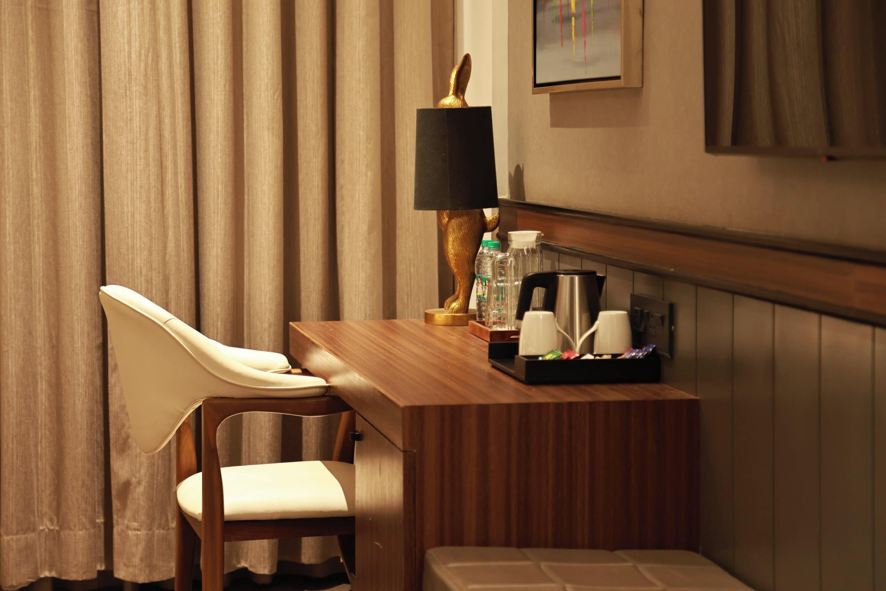 Habitación de hotel con escritorio de madera, una silla, una lámpara, una cafetera y una mesita con tazas y una botella.