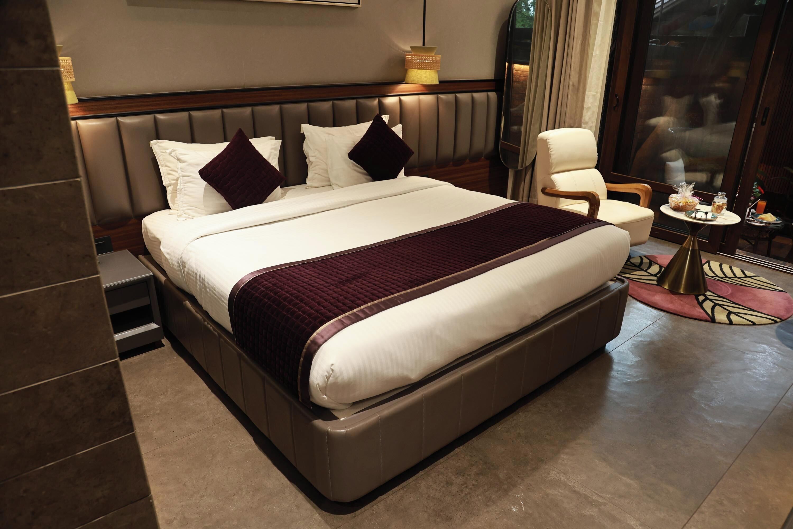 A hotel room with a large bed, a chair, a small table with a drink, and a window with curtains.