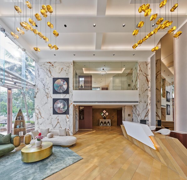 A spacious living area with a high ceiling, marble walls, and a wooden floor. There are hanging light fixtures and a large window providing natural light. - Kamah, Goa (Anjuna)
