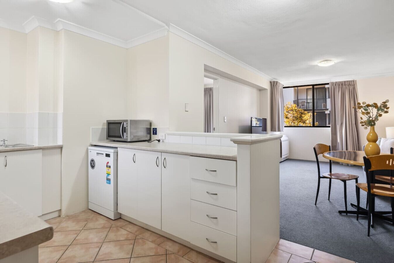 A compact kitchen with white cabinets, a microwave, and a small refrigerator. There is a dining area with a table and chairs, and a window with curtains.