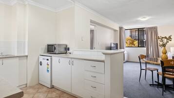 A compact kitchen with white cabinets, a microwave, and a small refrigerator. There is a dining area with a table and chairs, and a window with curtains.