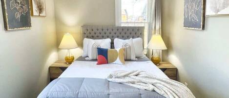 A neatly made bed with a grey headboard, two bedside lamps, and a window with curtains.