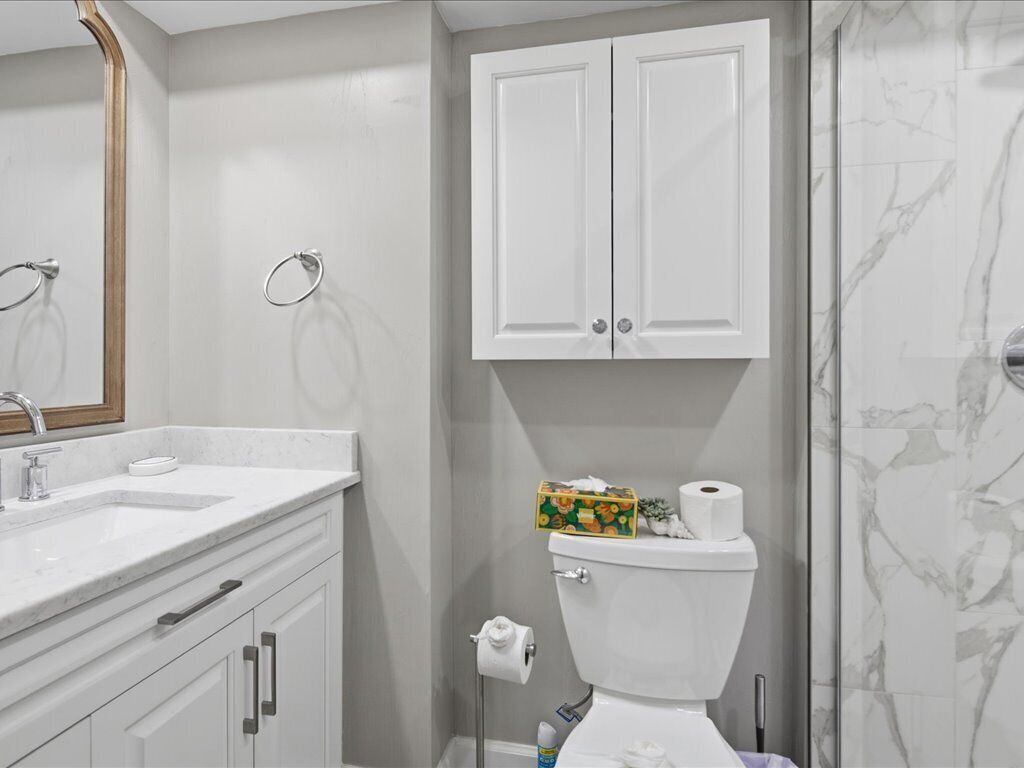 A bathroom with a white sink, mirror, two cabinets, toilet, and a shower area.