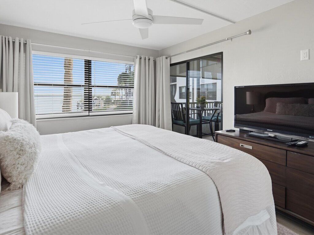 Un dormitorio con cama, televisión, escritorio y balcón con vista a edificios.