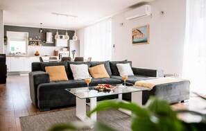Een moderne woonkamer met een donkergrijze bank, een glazen salontafel en een keuken op de achtergrond.