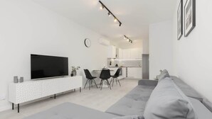 Una sala de estar moderna con un televisor de pantalla plana grande, un sofá gris, un comedor con sillas y una cocina al fondo.