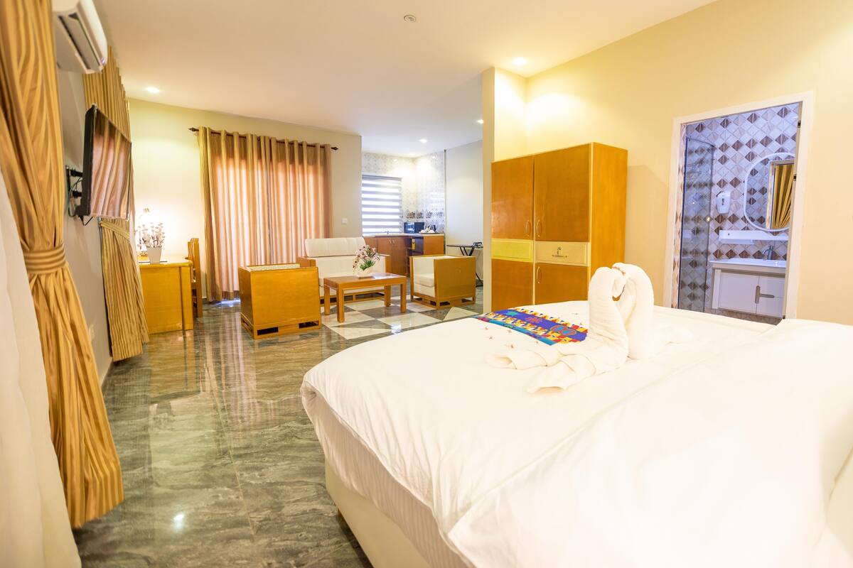 A hotel room with a large bed, a desk, a chair, a wardrobe, and a bathroom visible through a glass door.