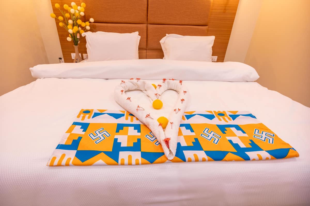 A neatly made bed with white linens, a heart-shaped towel with a geometric pattern, and a vase of yellow flowers on the nightstand.