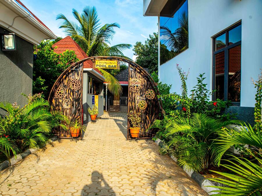 Un sendero conduce a un bar junto a la piscina, con un arco tropical, rodeado de plantas en macetas y palmeras.