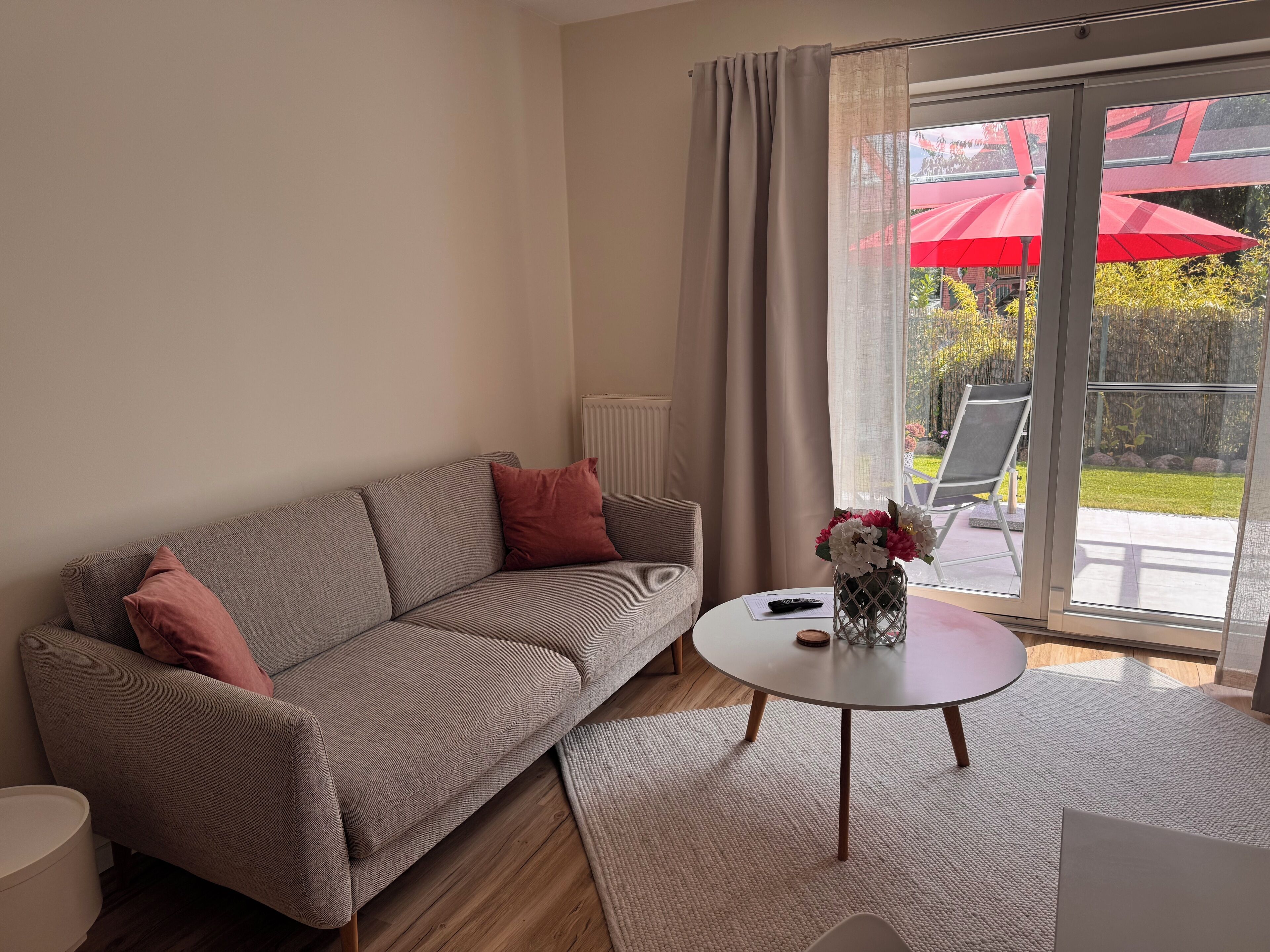 A modern living room with a grey sofa, a small round table, and a view of a garden through a sliding glass door.
