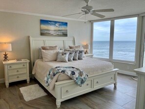 Une chambre dotée d’un grand lit, d’un ventilateur de plafond, d’une table de chevet avec une lampe et offrant une vue sur l’océan par le biais d’une grande fenêtre.