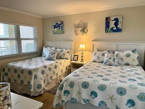 A bedroom with two beds, a bedside table with a lamp, and a window with blinds.