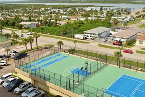 A tennis court with a net, surrounded by a fence, and adjacent to a parking area with several cars.