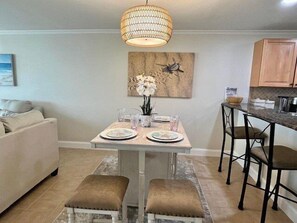 A dining area with a table set for four, a chandelier, a painting, and a kitchen area with a bar. - Shores Club #602 - Daytona Beach Oceanfront Vacations, Inc (Daytona Beach Shores)