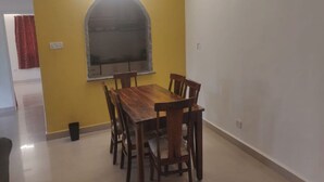 A dining area with a wooden table and chairs, a window with a decorative arch, and a red curtain on the left. - Signum Court (Calangute)