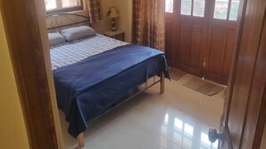 A bedroom with a bed, a nightstand, a lamp, and a wooden door. - Signum Court (Calangute)