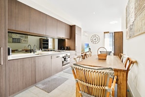 A modern kitchen with wooden cabinets, a dining area with a wooden table and wicker chairs, and a wall clock.