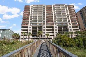 A multi-story residential building with balconies, a wooden boardwalk in front, and surrounding greenery.