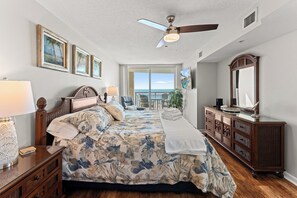A bedroom with a bed, a ceiling fan, a dresser, a TV, and a large window with a view.